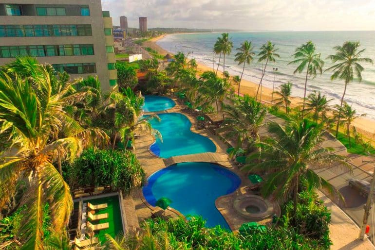 Imagem de uma piscina em frente a praia com o hotel ao fundo da piscina.