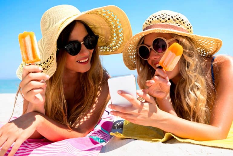 Mulheres deitadas na praia tomando sorvete e olhando para o celular