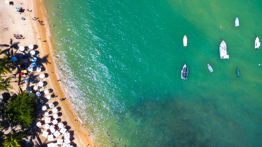 Foto tirada nos céus da praia de Arraial d'Ajuda com embarcações na água e pessoas na praia