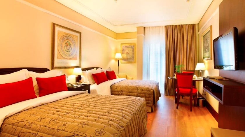 Imagem de um quarto de hotel com duas camas de casais com uma tv a frente, mesa e cadeiras.
