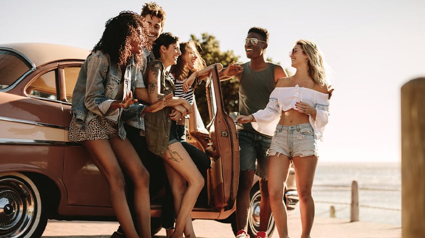 Grupo de amigos conversando em frente à uma carro, com a praia de cenário ao fundo