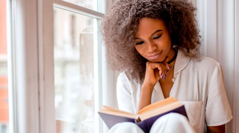 Mulher, próxima à uma janela, lendo um livro